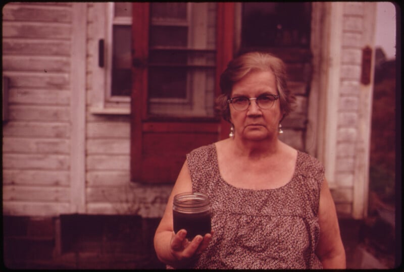 An older woman with glasses stands in front of a weathered white house, holding a jar filled with dark liquid. She wears a sleeveless patterned dress and has a serious expression.