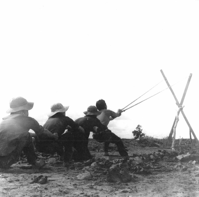 Four children sit outdoors on rough ground, watching as one child uses a large, Y-shaped slingshot made of wooden poles, aiming at something off-frame under a bright sky.