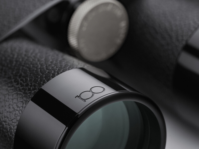 Close-up of a black binocular lens with "100" engraved on the rim, featuring textured grips and a metal focusing knob in the background.
