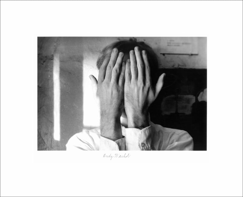A person in a white shirt stands facing the camera with their hands covering their face. The photo is black and white, with a blurred background and a faint signature beneath the image.