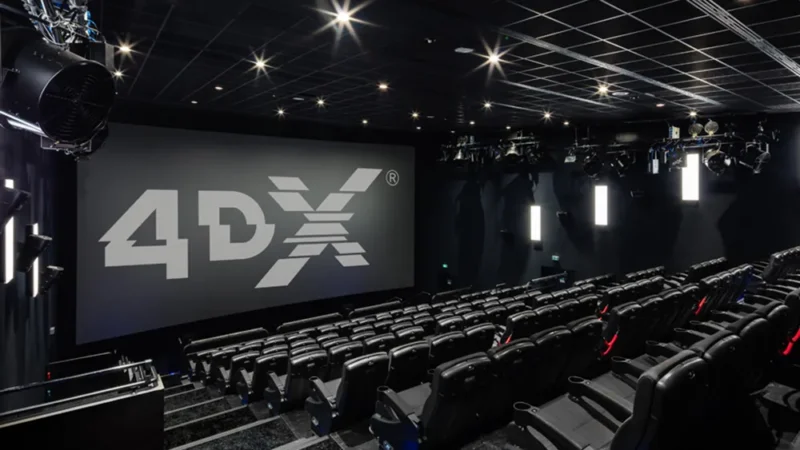 A modern 4DX movie theater with black seats and a large screen displaying the 4DX logo. The room has ceiling lights, motion equipment, and a dark, immersive atmosphere.