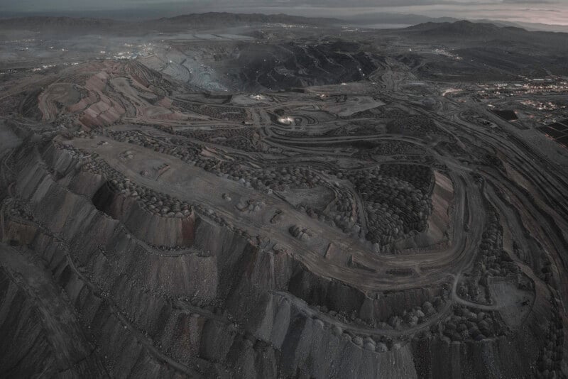 Aerial view of a large open-pit mine with terraced, winding roads and exposed earth layers, surrounded by mountains and a hazy sky in the background. The landscape is rugged and industrial.