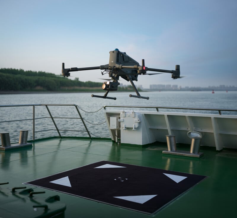 A drone hovers above a marked landing pad on the deck of a boat, with calm water and a distant shoreline visible in the background under a clear sky.