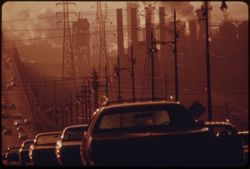Cars are stuck in traffic on a road lined with power lines, with industrial smokestacks in the background releasing thick smoke into a hazy, reddish sky.