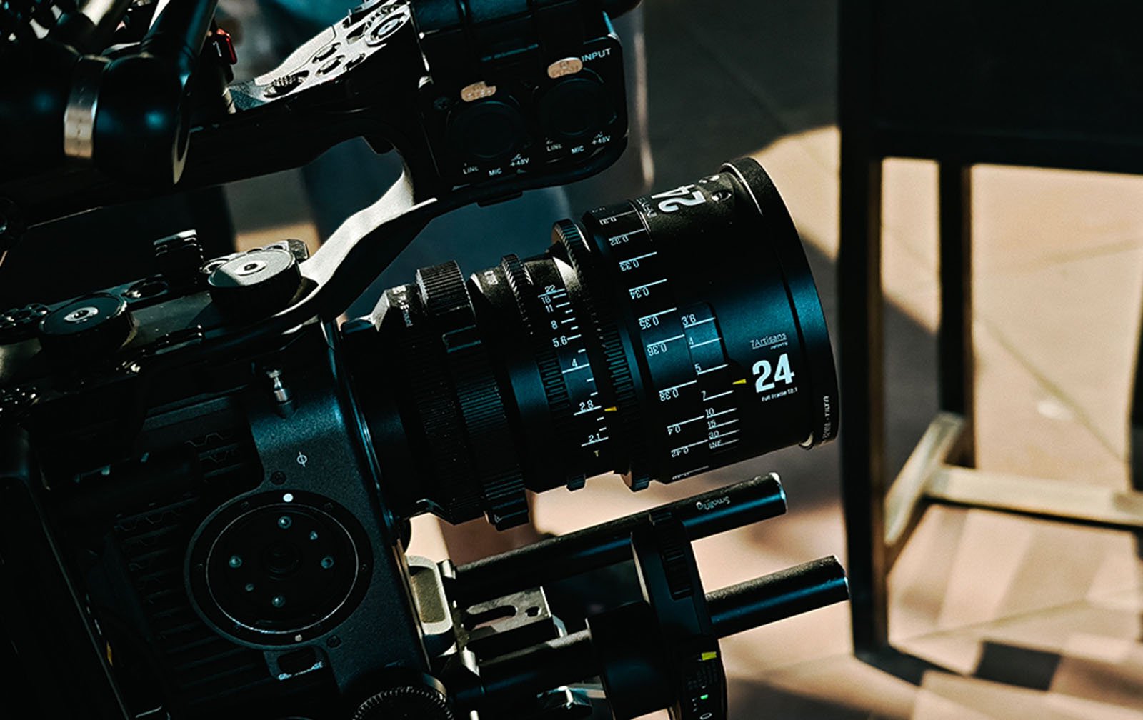 Close-up of a professional film camera with a 24mm lens, showing detailed focus and aperture markings, set up on a tripod with control handles visible.