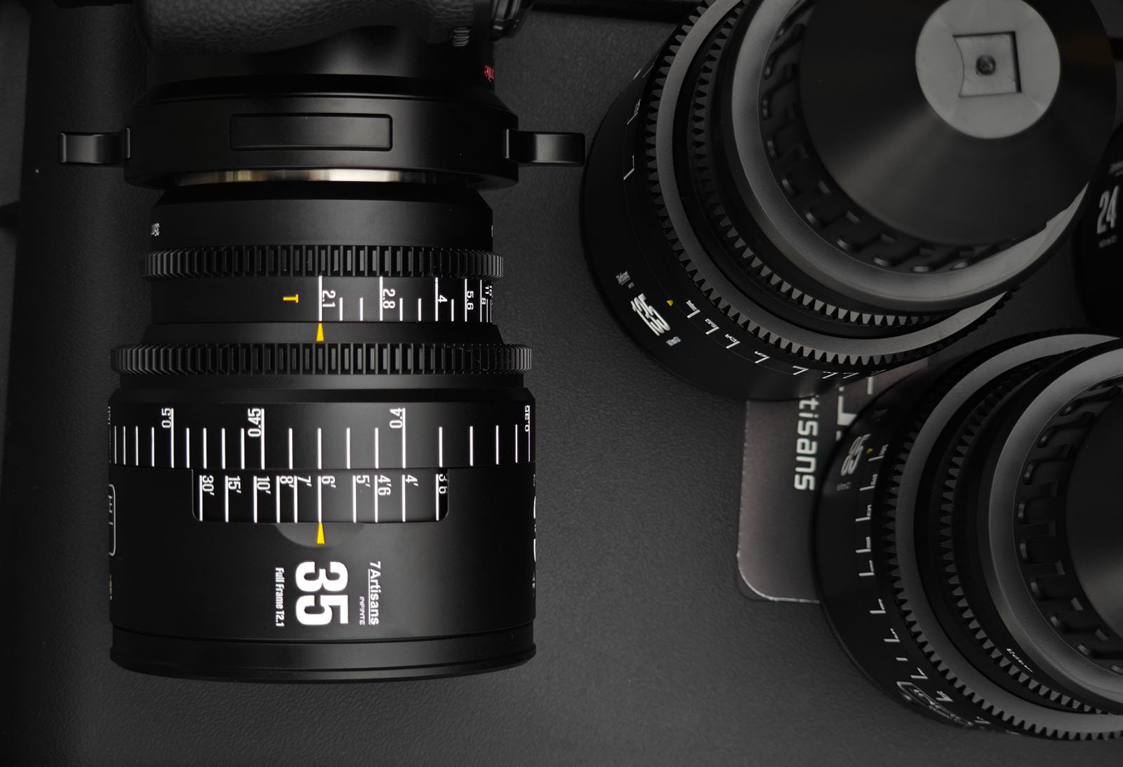 Close-up of a 35mm cinema camera lens mounted on a camera, with additional camera lenses placed nearby on a black surface. The image highlights detailed focus and aperture markings on the lenses.