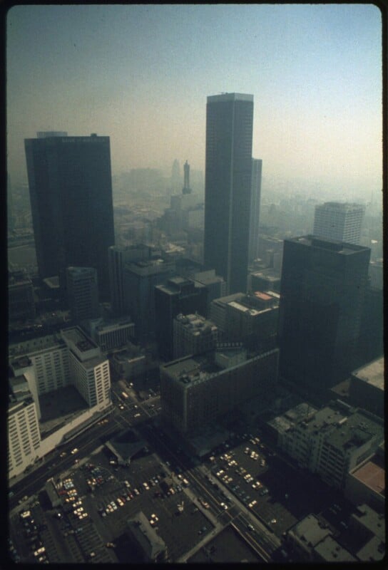 Aerial view of a city with tall skyscrapers and dense smog, reducing visibility. The streets below show cars in parking lots and on the roads, with buildings clustered closely together.