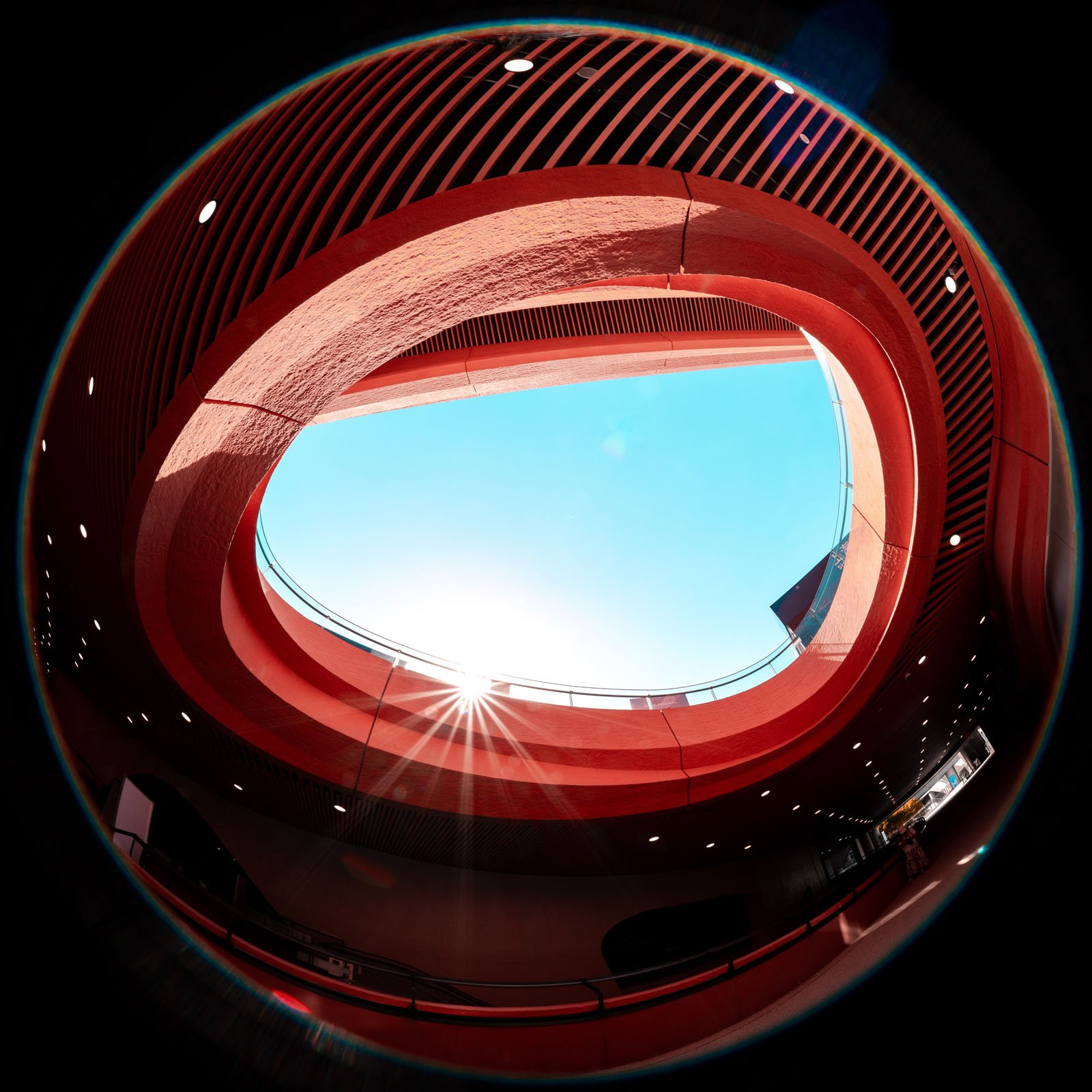 منظره ای از Fisheye که از طریق یک دهانه بیضی شکل در یک ساختمان قرمز مدرن که در آن نور خورشید جریان دارد و یک آسمان آبی روشن در بالا مشاهده می شود ، نگاه می کند.