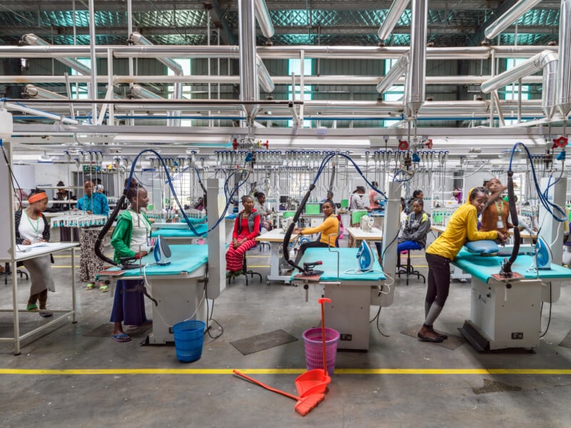 A group of workers, mostly women, operate ironing and pressing stations in a brightly lit garment factory with industrial equipment and pipes overhead.