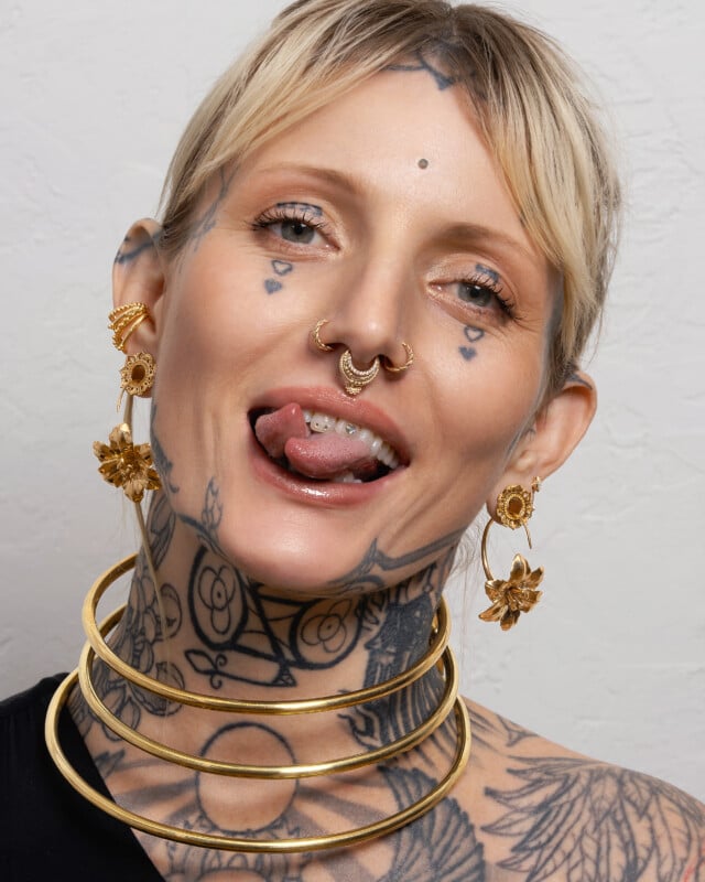 A person with blonde hair, facial and neck tattoos, wearing gold earrings, nose rings, and three large gold neck rings, sticks out their tongue and smiles. The background is a plain, light-colored wall.