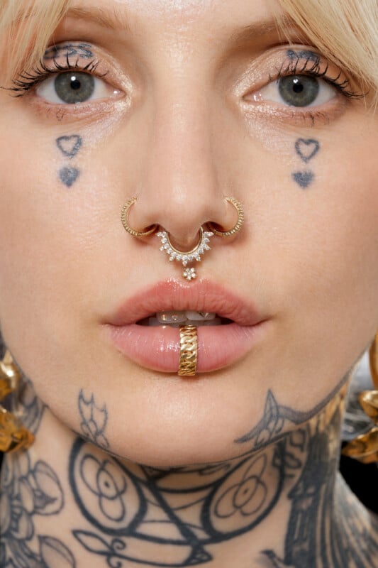 A close-up of a person with light eyes, heart-shaped face tattoos, gold and jeweled nose rings, a gold ring in their lower lip, and visible neck tattoos, with blonde hair framing their face.