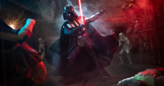 Darth Vader wields a red lightsaber in a dramatic, smoky scene with stormtroopers and other figures in the background, illuminated by red and blue lighting.