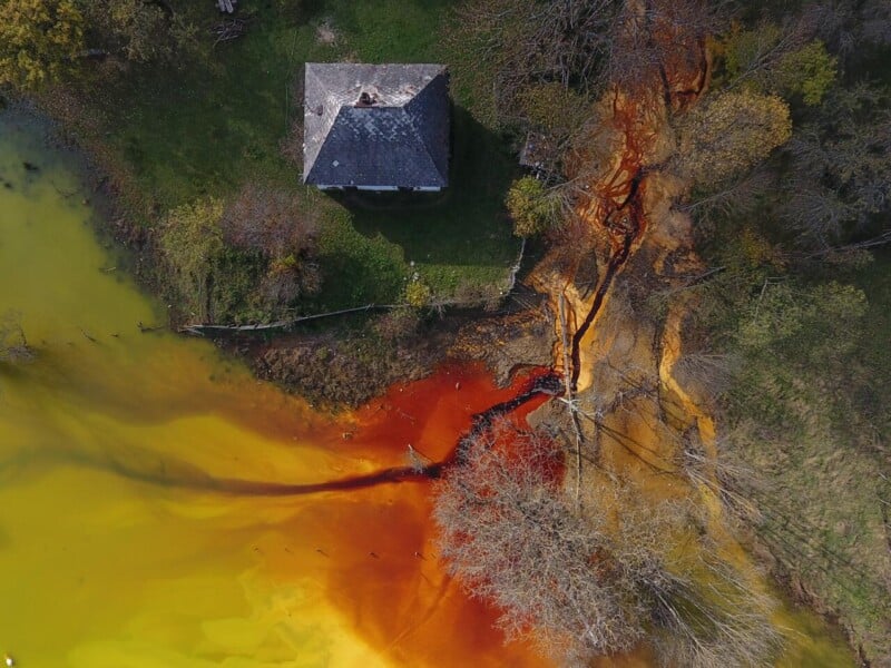 Aerial view of a small house near a landscape with yellowish water blending into vivid orange and red, possibly due to pollution, with some trees and sparse grass surrounding the area.