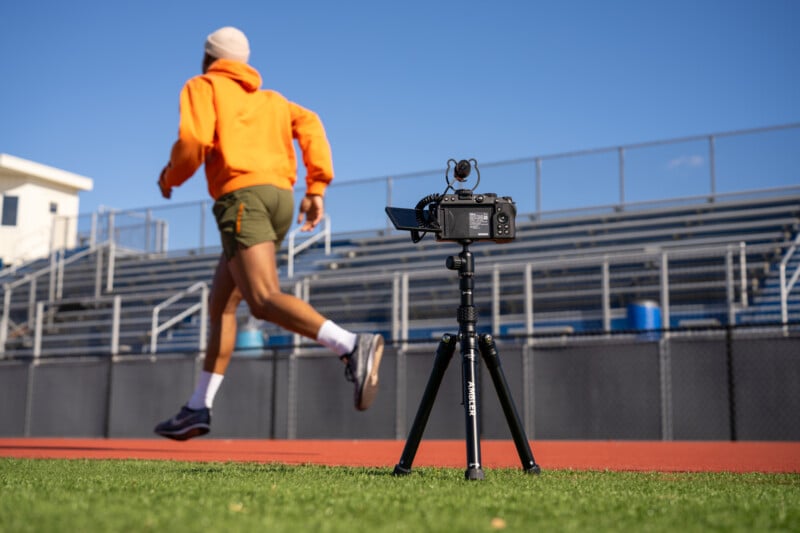 A camera on a tripod films a person in an orange hoodie and shorts running on a track beside empty bleachers under a clear blue sky.