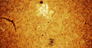 A high-resolution image of the Sun’s surface shows swirling patterns, a sunspot, a solar flare, and the silhouette of the International Space Station passing in front of the Sun. The scene is colored in vivid orange and yellow tones.