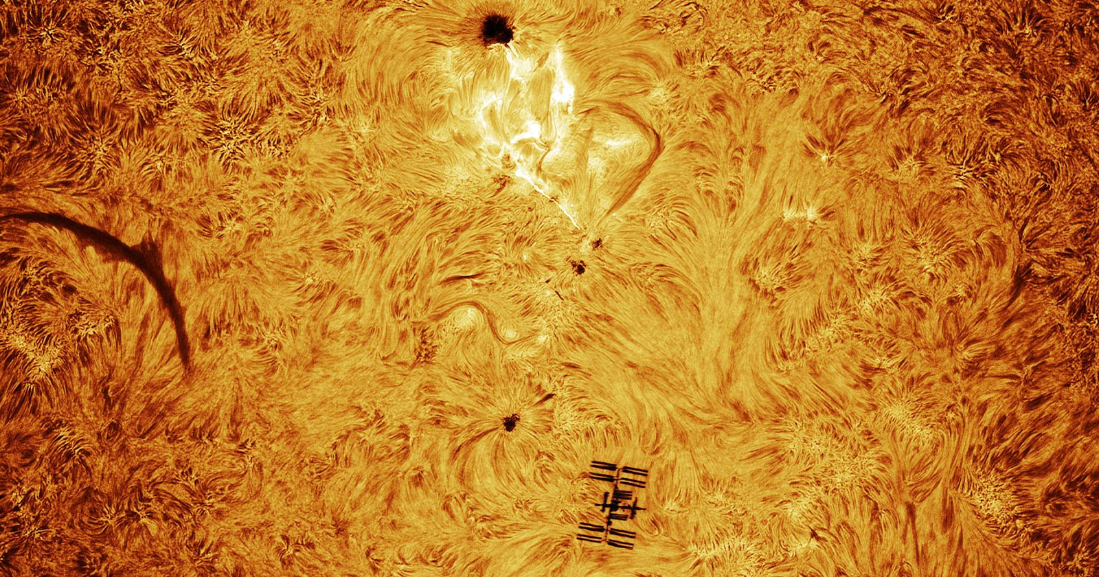 A high-resolution image of the Sun’s surface shows swirling patterns, a sunspot, a solar flare, and the silhouette of the International Space Station passing in front of the Sun. The scene is colored in vivid orange and yellow tones.