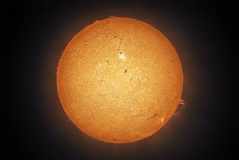 A detailed, close-up image of the Sun, showing its textured surface with bright, glowing orange and yellow colors, wispy solar prominences, and darker sunspots against a black background.