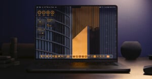 A laptop on a dark desk displays a desktop with orange and black geometric wallpaper, orange-themed icons, and widgets showing system stats and weather. The background includes modern building structures.