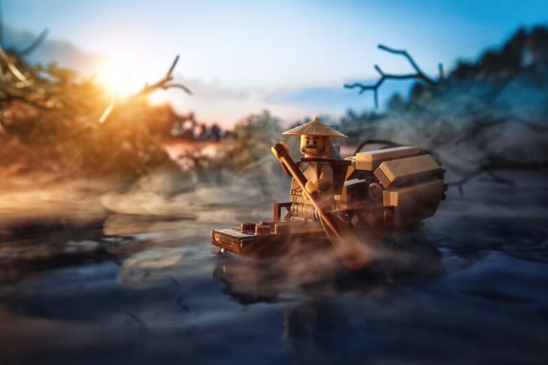 A LEGO figure wearing a conical hat rows a small wooden boat through misty water at sunrise, surrounded by blurred branches and soft morning light.