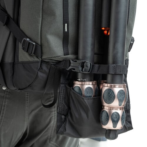 A close-up of a black backpack with a tripod secured in a side pocket, showing the tripod’s adjustable legs and locking mechanisms. The person wearing the backpack is partially visible, dressed in dark clothing.