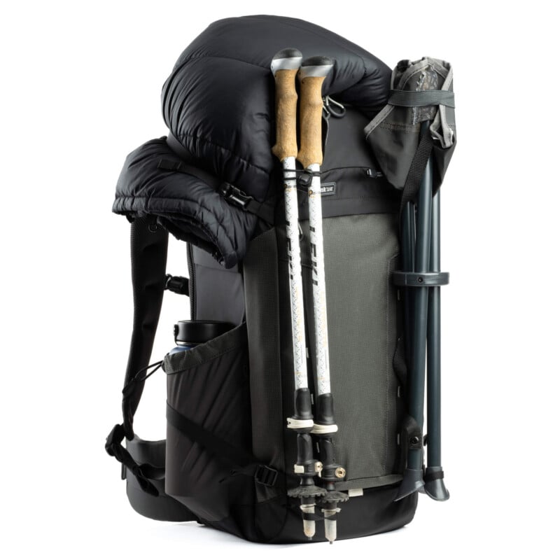 A fully loaded hiking backpack with a black jacket secured on top, two trekking poles attached to the side, a water bottle in the side pocket, and a folded tripod fastened on the opposite side.