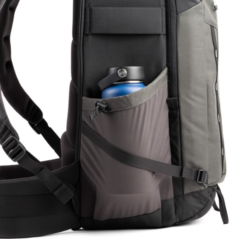 A close-up of a backpack's side pocket holding a blue water bottle with a black lid. The backpack has padded straps and a mix of grey and black fabric.