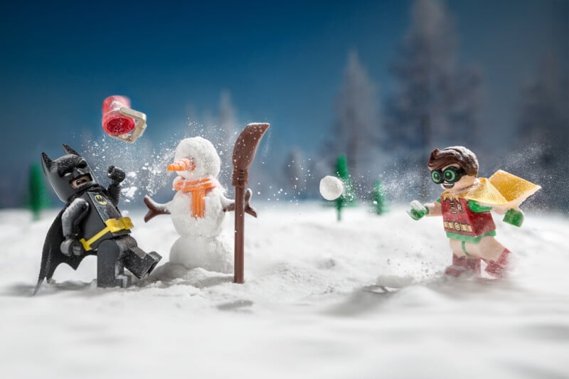 Lego Batman is hit by a snowball thrown by Lego Robin in a snowy landscape, with a snowman and winter trees in the background.