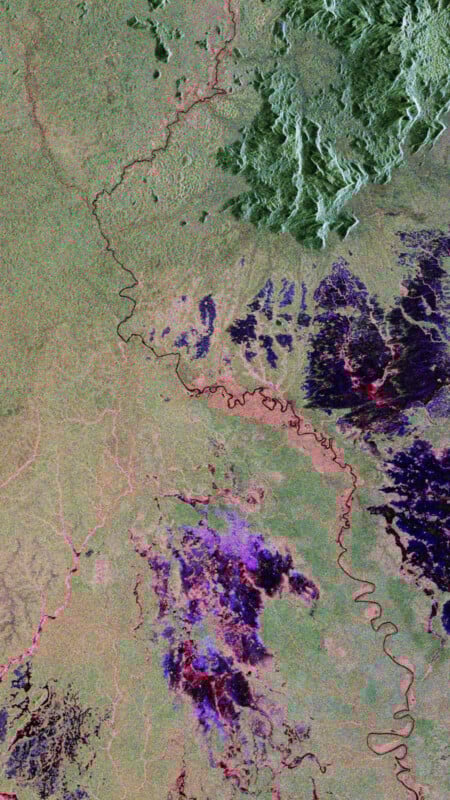 Satellite image showing a winding river cutting through varied terrain with patches of green, purple, and pink, indicating different vegetation and land types. Dense forested areas are seen in the upper right and central regions.