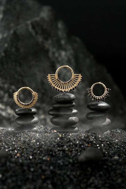Three ornate gold rings are displayed on stacks of smooth black stones, with a misty, dark rock background creating a dramatic and elegant atmosphere.