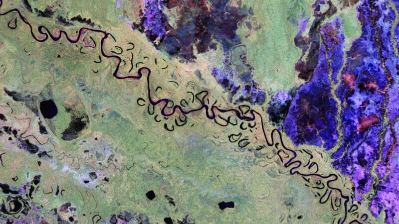 A satellite image showing a winding river with many bends and loops through green and purple terrain, surrounded by patches of blue and black, indicating varied landscape features.