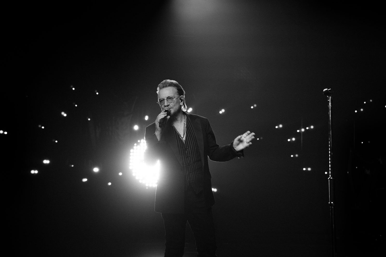 A person in a suit sings on stage in dramatic lighting, holding a microphone with bright lights and an illuminated audience in the background, creating a moody concert atmosphere.