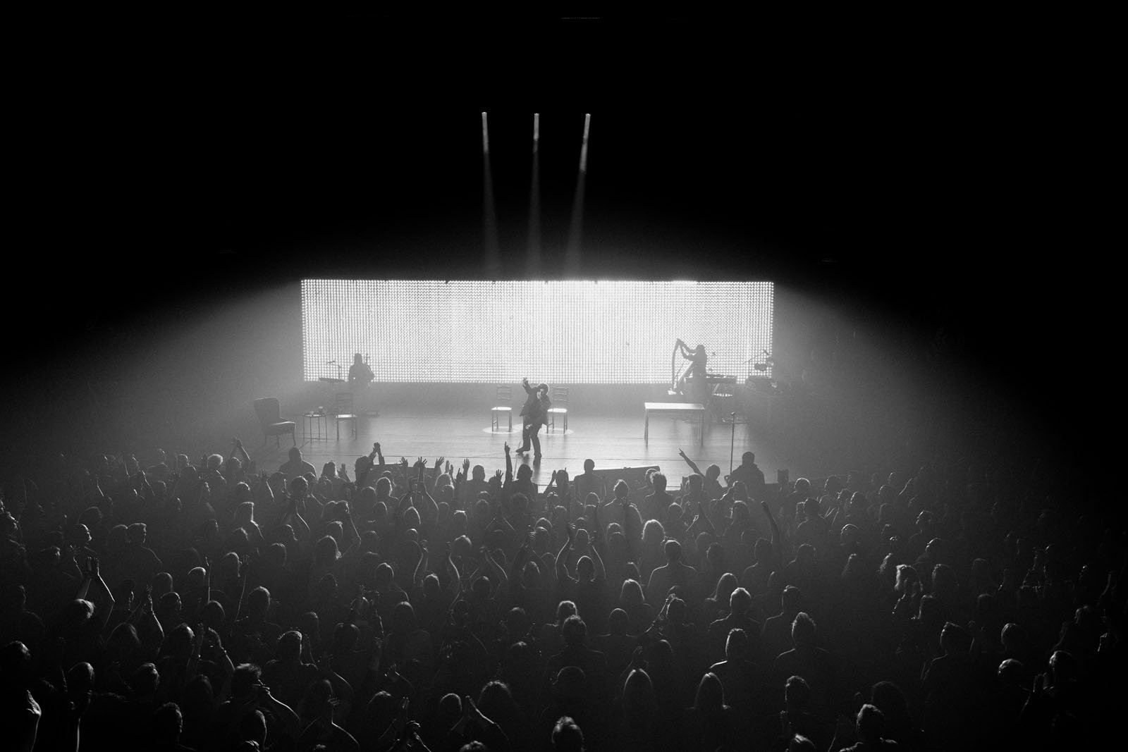 A black and white photo of a concert stage with performers silhouetted against a bright, backlit screen. A large, enthusiastic audience fills the foreground, facing the stage.