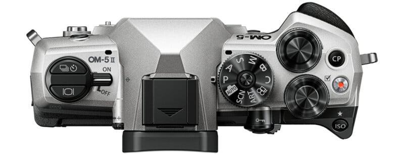 Top view of a silver Olympus OM-D camera showing control dials, buttons, power switch, shutter button, mode dial, hot shoe, and textured grip on the right side.
