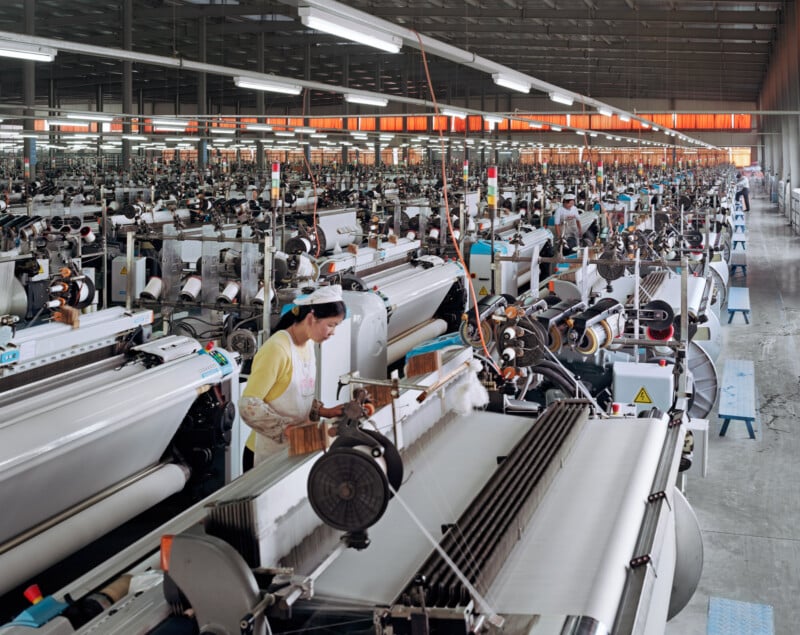 A large textile factory with rows of weaving machines; a worker in a yellow shirt operates one machine in the foreground under bright overhead lights, with many more workers and equipment visible in the background.
