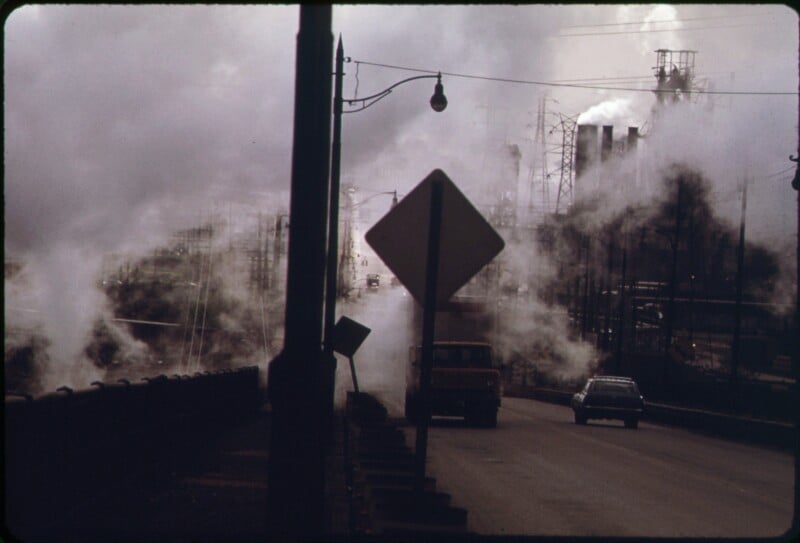 Cars drive on an industrial road enveloped in thick smoke or steam, with large factories and power lines in the background and a road sign visible in the center of the image.