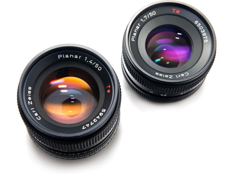 Two Carl Zeiss camera lenses, labeled "Planar 1.4/50" and "Planar 1.7/50," are placed on a white surface with colorful reflections visible on their glass elements.