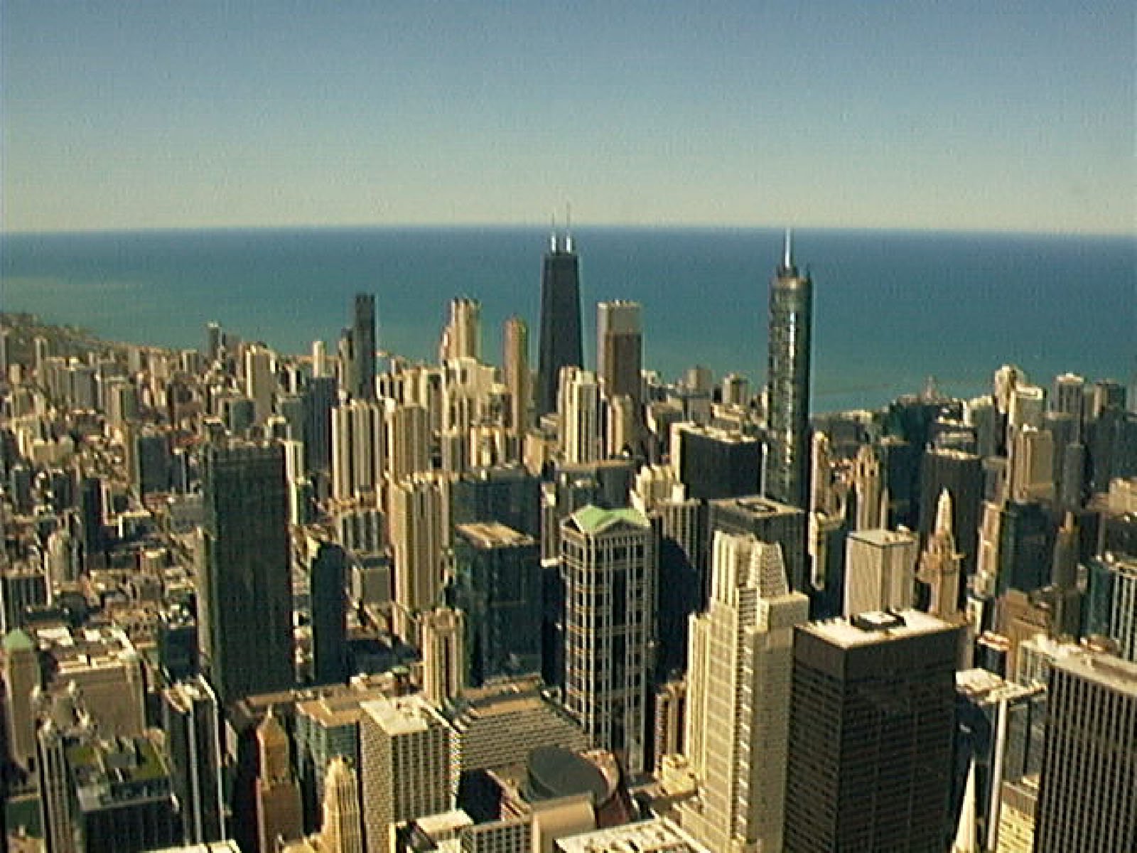 Aerial view of a city skyline with many tall skyscrapers and buildings near a large body of water under a clear blue sky.