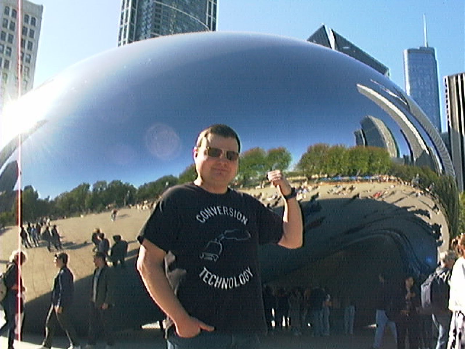 A man wearing sunglasses and a "Conversion Technology" T-shirt poses with a thumbs-up in front of Chicago’s Cloud Gate (The Bean) sculpture, with city buildings and people reflected in the shiny surface.