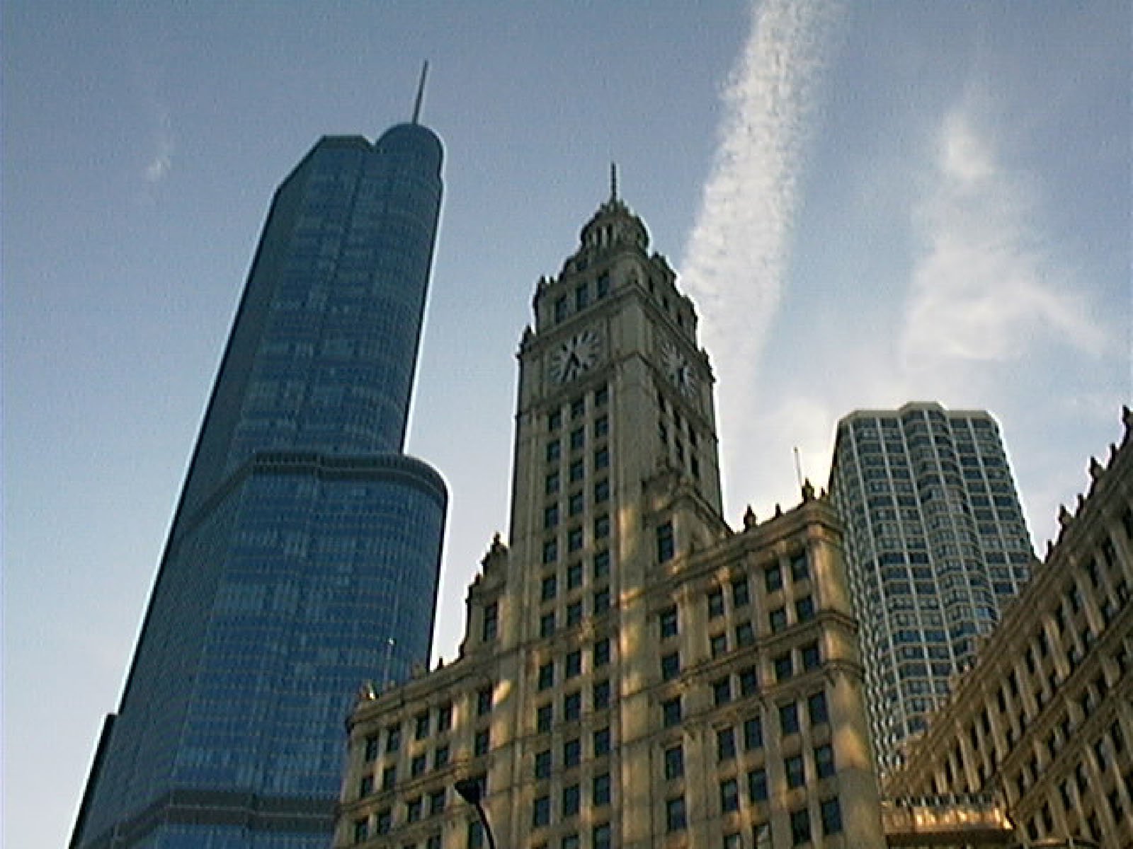 Three tall buildings rise into a blue sky; the central one has a clock tower, flanked by a modern glass skyscraper and a rounded building. Sunlight and a streak of cloud are visible.