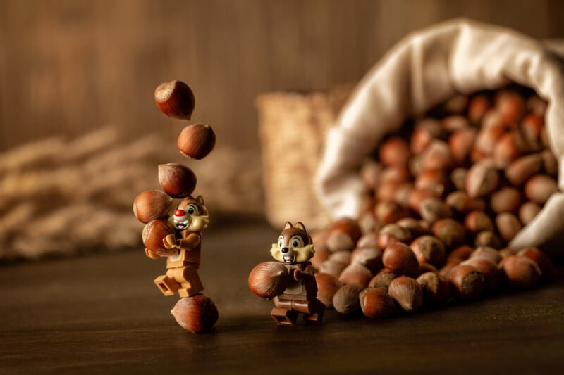 Two LEGO chipmunk figurines hold and balance hazelnuts, with more nuts spilling from a woven basket in the background. The scene is playful and set on a dark wooden surface.