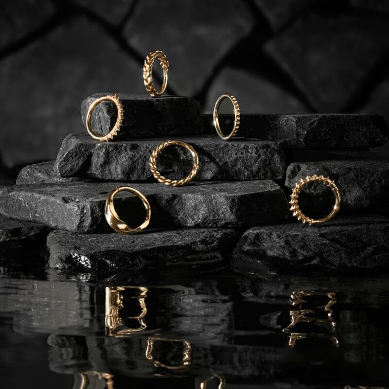 Six gold rings with various textured designs are displayed on stacked black stones, with their reflections visible in the dark water below, creating a dramatic and elegant scene.