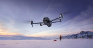 A drone hovers in the foreground above a snowy landscape at sunset, while a person stands on the snow below near equipment with mountains visible in the background.