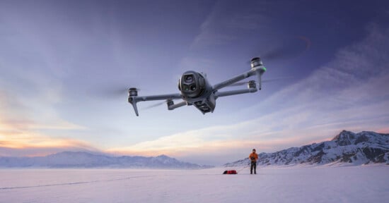 A drone hovers in the foreground above a snowy landscape at sunset, while a person stands on the snow below near equipment with mountains visible in the background.