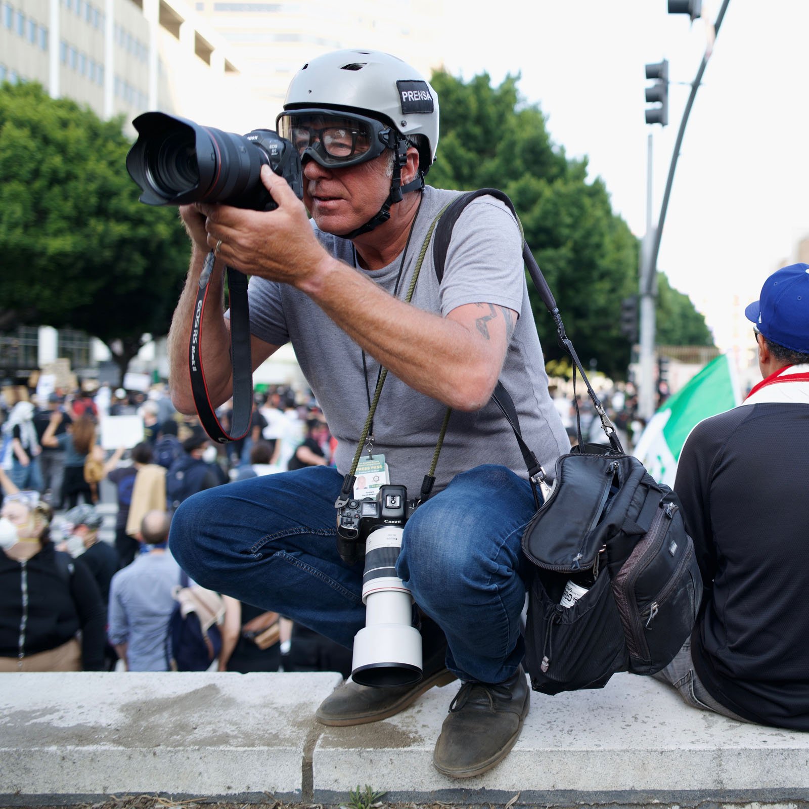 A man wearing a helmet and sunglasses crouches on a concrete ledge, holding a camera and aiming to take a photo at a busy outdoor event with a crowd in the background.