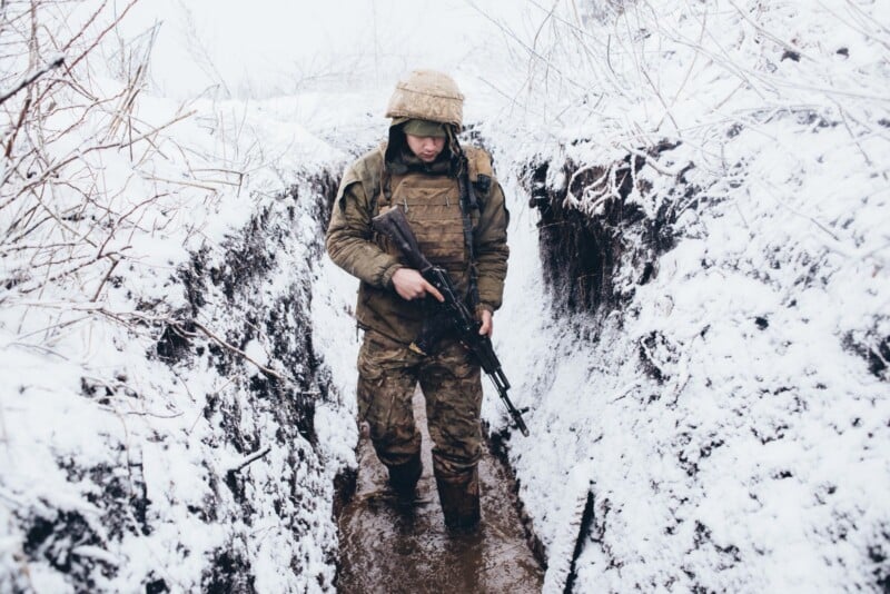 A soldier in camouflage gear and helmet stands in a muddy, snow-covered trench, holding a rifle and looking down, with bare branches visible on both sides.