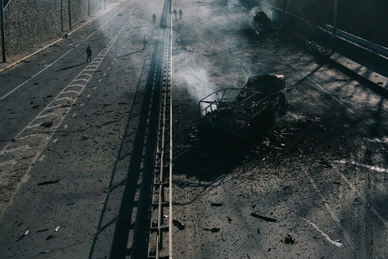 A burned-out vehicle sits on a debris-strewn, smoke-filled road divided by a fence. A few people walk in the distance, and shadows cast by the railing and smoke suggest a recent destructive event.