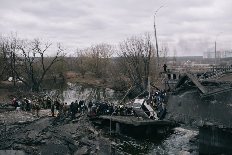 A large group of people crosses the debris of a destroyed bridge over a river, surrounded by leafless trees and cloudy skies, with damaged infrastructure in the background.
