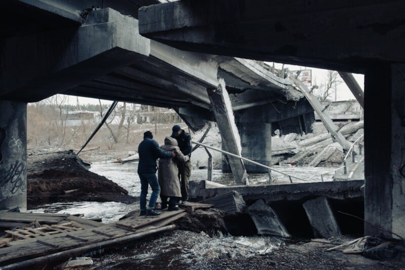 Three people help each other navigate rubble and debris beneath a destroyed concrete bridge, with broken sections hanging over a river. The scene appears cold and somber, suggesting recent destruction or conflict.