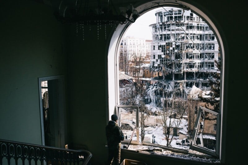 A person in military clothing stands by a broken window inside a damaged building, looking out at another heavily destroyed building across a snowy courtyard. Daylight streams through the large arched window.