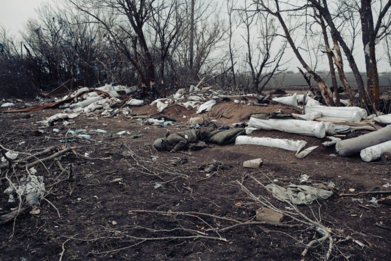 A desolate, muddy area with scattered debris, fallen branches, plastic-wrapped items, and what appears to be a soldier lying on the ground among leafless trees under an overcast sky.